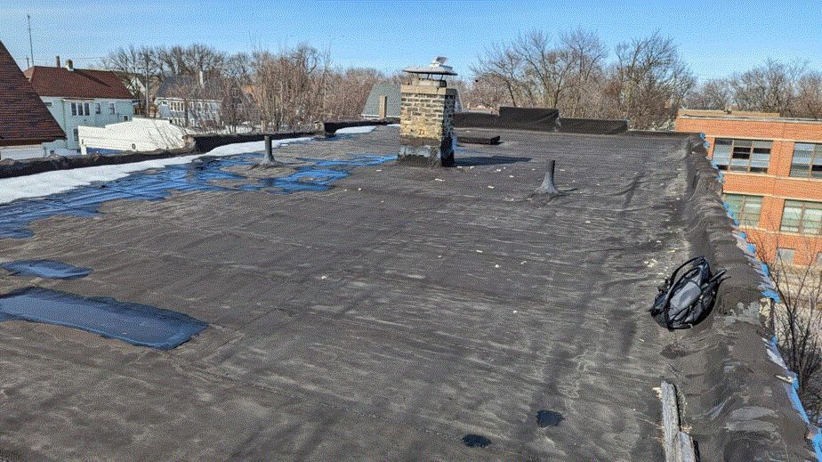 Flat rooftop with black tar paper, some patches, puddles, vents, and a brick chimney; snow remnants on the left edge, buildings and bare trees in the background under a clear blue sky.