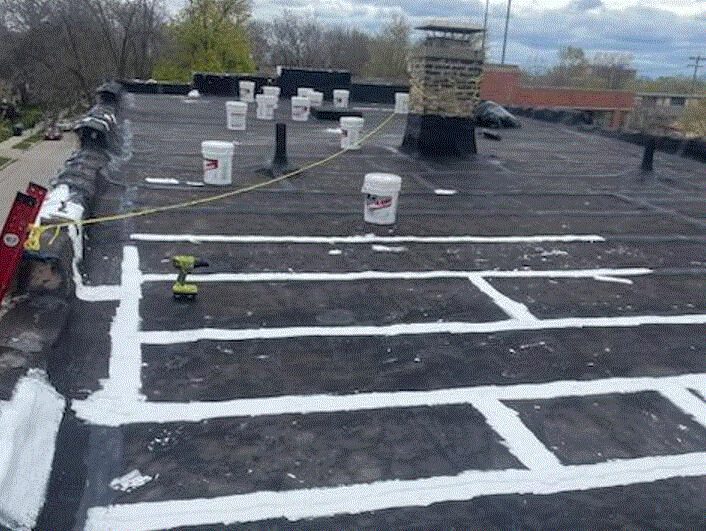 A flat rooftop with several buckets, a cordless drill, and white sealant applied in straight lines, forming a grid pattern. A yellow rope is stretched across the roof near a brick chimney. Trees and buildings are visible in the background.