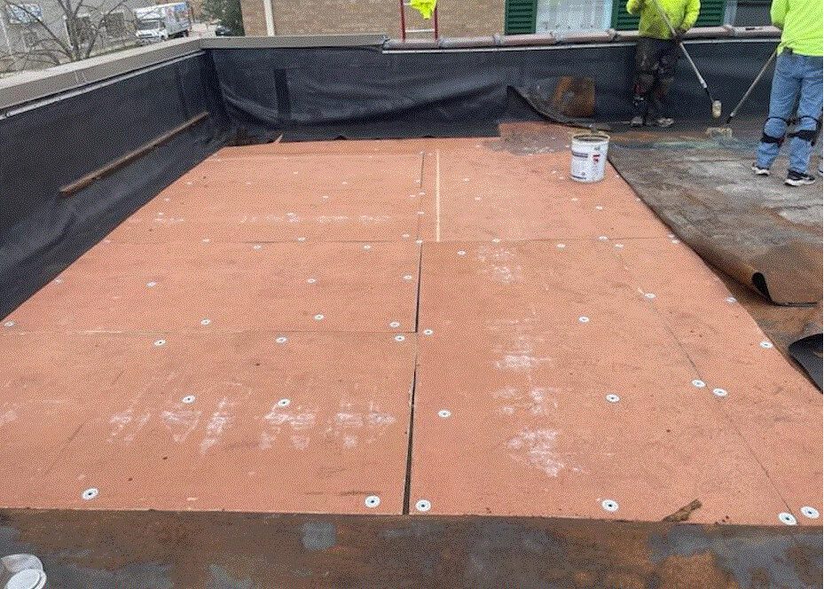 Several plywood boards, fastened with screws and washers, cover part of a flat rooftop. Two workers in safety gear stand near the edge, with tools and a can of roofing material visible.