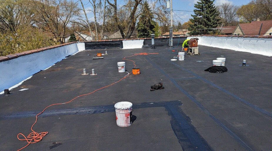 A flat rooftop is being worked on, with black roofing material, white paint buckets, tools, and extension cords scattered around. Two workers are visible in the distance near the roof’s edge on a sunny day.