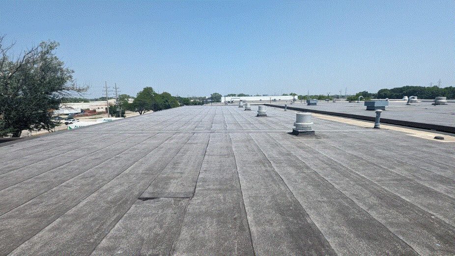 A flat, gray industrial rooftop with vents and pipes, stretching into the distance under a clear blue sky. Trees and buildings are visible on the horizon.