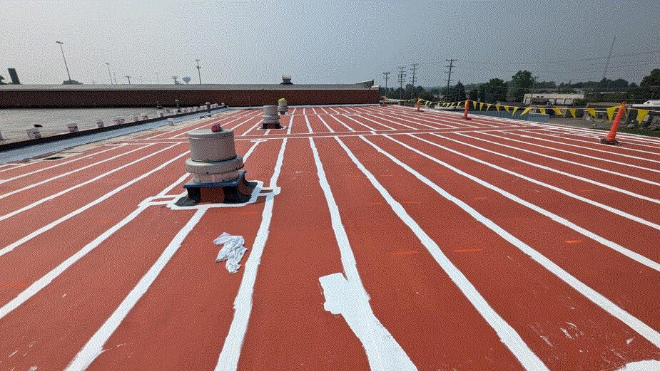 A flat rooftop coated in red with evenly spaced white stripes, featuring several vents and safety cones. Yellow caution tape is strung along the right side, and industrial structures are visible in the background.