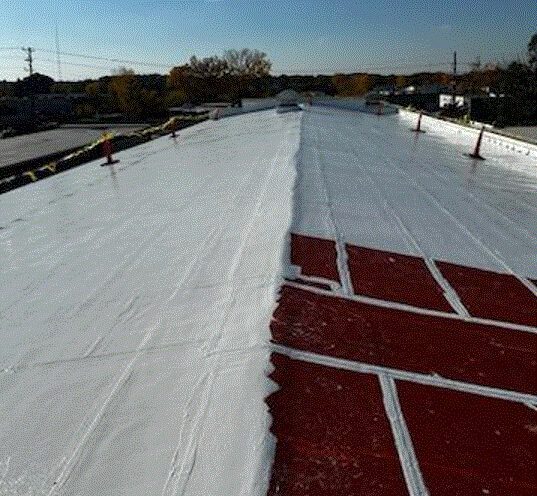 A flat commercial roof partially coated with white sealant, with some red primer or undercoating still visible. Safety cones and caution tape line the roof edges. Trees and buildings are in the background under a clear sky.