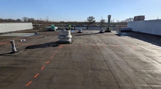 A flat commercial rooftop with various vents, pipes, and HVAC units under a clear blue sky. Orange dashed lines are marked on the roof, and a few workers are visible in the distance.