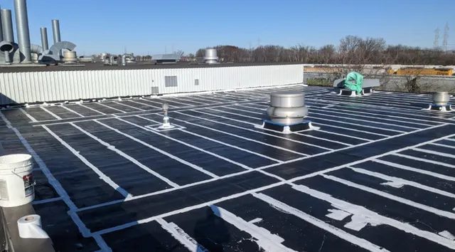 A flat commercial roof with black waterproofing membrane and white sealant lines applied in a grid pattern, ventilation units, pipes, and industrial buildings visible in the background under a clear sky.
