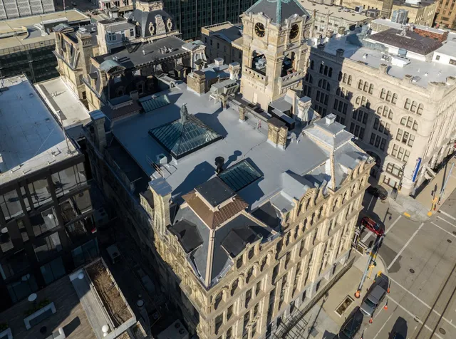 Aerial view of a historic stone building with a clock tower, surrounded by modern city structures and streets with light traffic under clear skies.