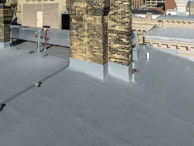 A flat commercial rooftop with a smooth, gray waterproof membrane, brick structures, and vents, set against a cityscape background with various building facades.