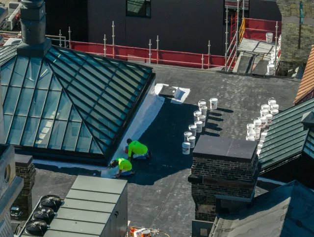 Two workers in bright yellow shirts apply material to a flat rooftop, surrounded by several white buckets. The roof includes a large skylight, brick structures, and adjacent construction scaffolding.