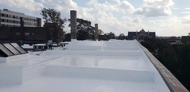 A flat white rooftop with raised sections, solar panels, and chimneys, overlooking trees and various buildings under a partly cloudy sky.