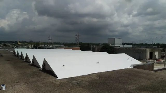 A row of white, triangular-roofed industrial buildings is seen under a cloudy sky, with urban structures and power lines visible in the background.