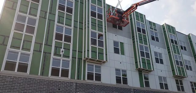 A construction worker in a lift works on the exterior of a multi-story building with green insulation panels, white siding, and a brick base. The sky is partly cloudy.