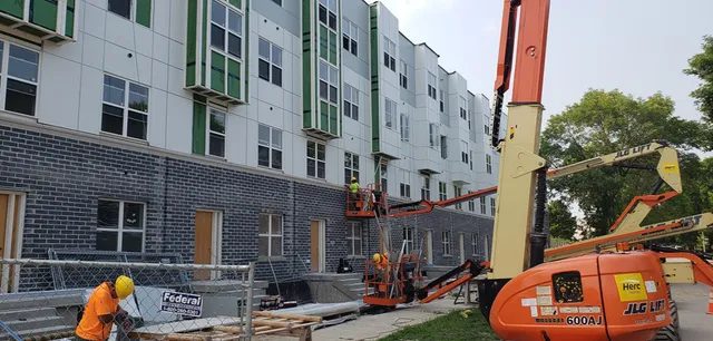 Construction workers in safety gear operate lifts and work on the exterior of a multi-story building under construction. The building has black brickwork on the lower level and white and green paneling above.