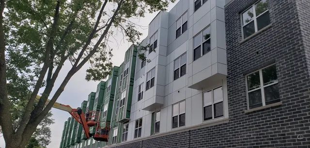 A construction lift is raised beside a multi-story building with gray brick and green siding, where workers appear to be working on the exterior. A large tree is in the foreground.