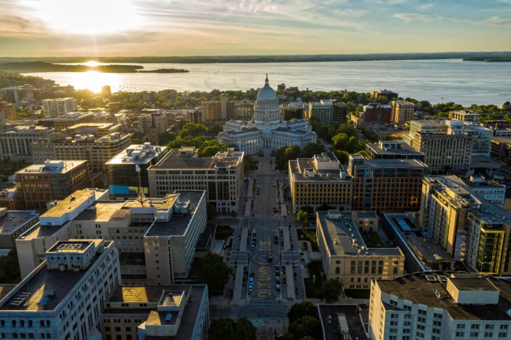 Aerial view of a city with a domed capitol building at the center, surrounded by office buildings and trees, with a large lake and sunset in the background.