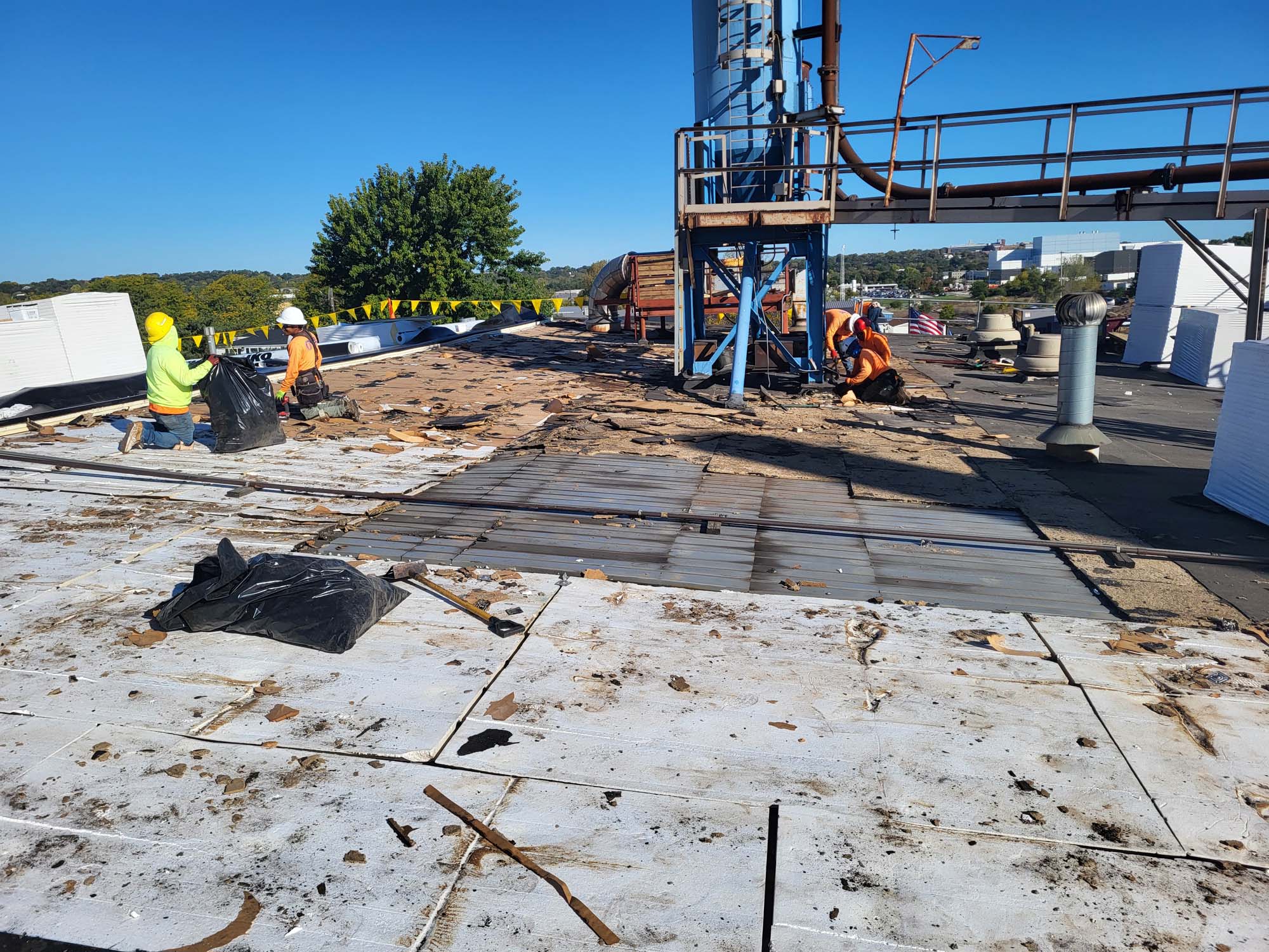 Several construction workers in safety vests and helmets work on a flat rooftop, removing old materials and debris under a clear blue sky, with construction equipment and trees in the background.