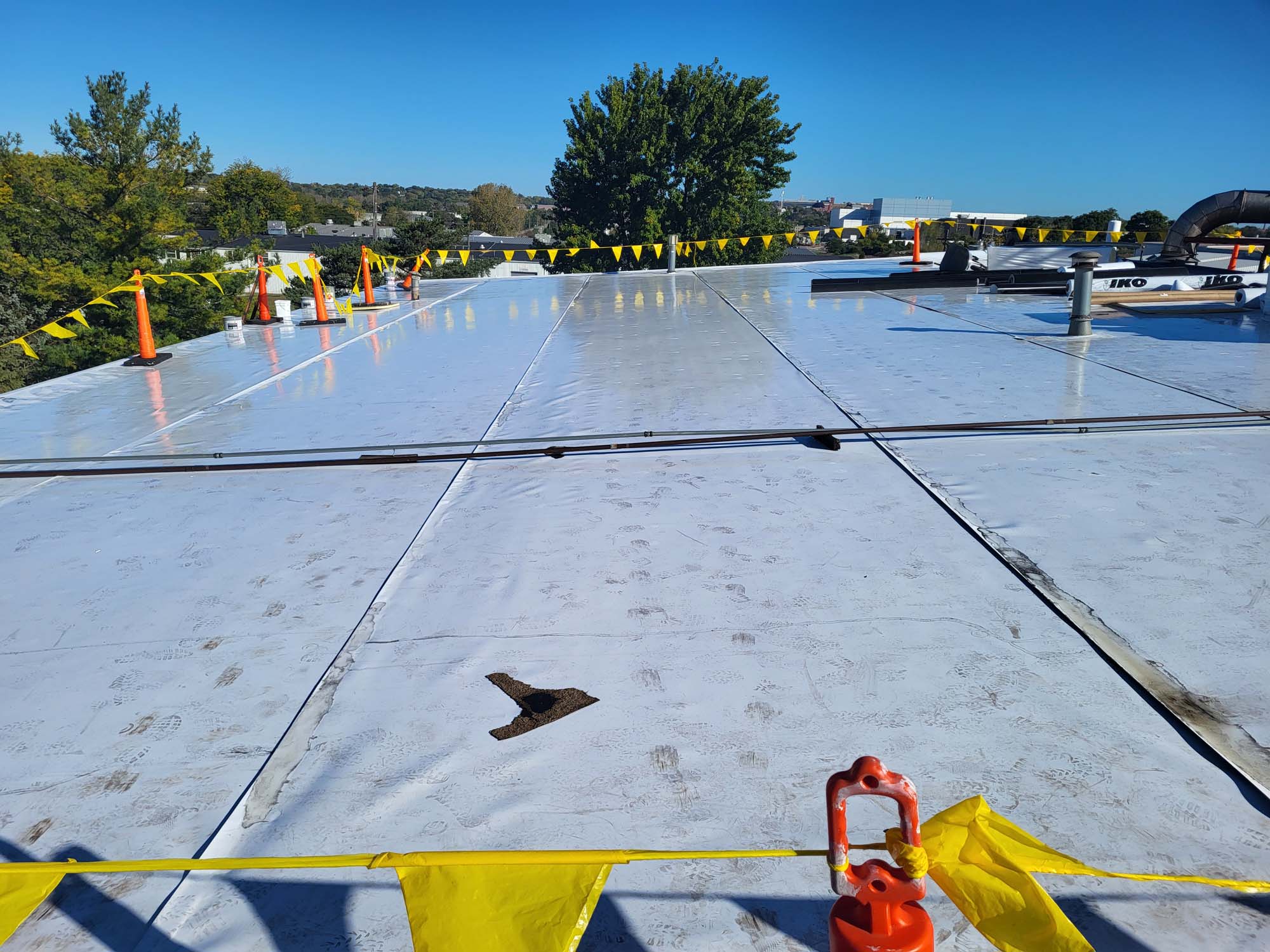 Flat commercial roof with a white reflective surface, bordered by yellow caution tape and orange cones. There are a few rooftop units and vents, and trees are visible in the background under a clear blue sky.