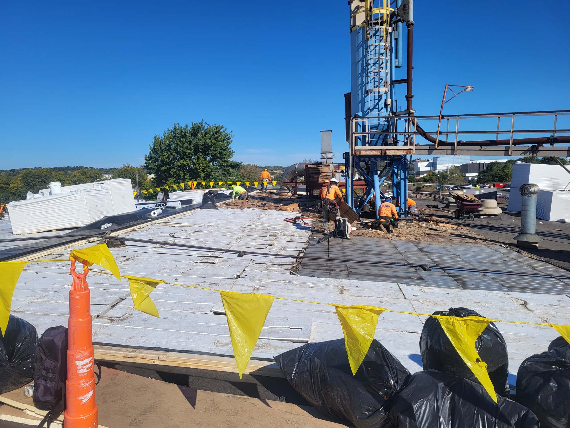 Construction workers wearing safety gear work on a rooftop, surrounded by yellow warning flags, black trash bags, insulation materials, and machinery on a clear, sunny day. Trees and buildings are visible in the background.
