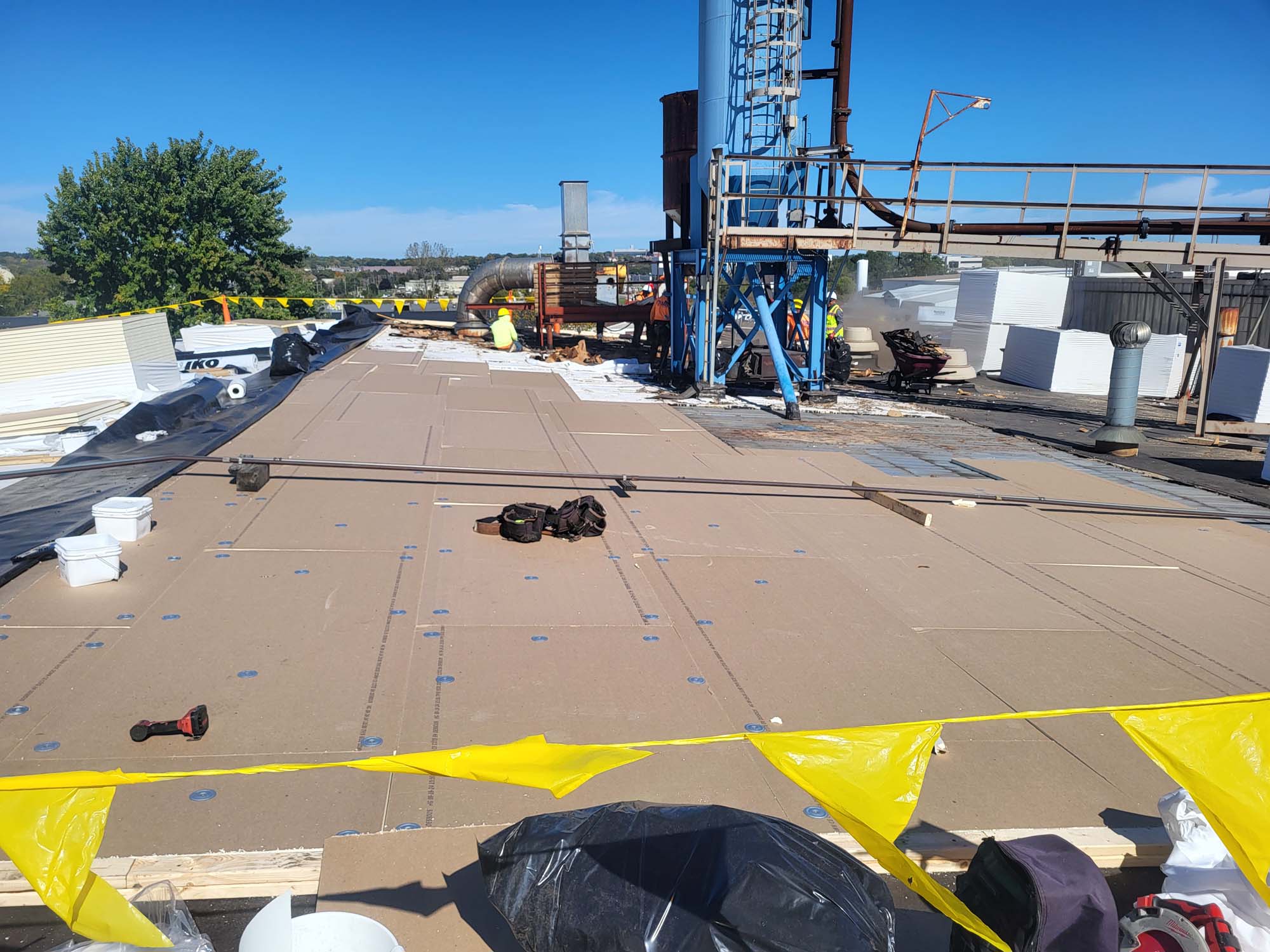 Rooftop construction site with insulation boards being installed, tools and materials scattered around, yellow caution tape in foreground, and industrial equipment in the background under a clear blue sky.