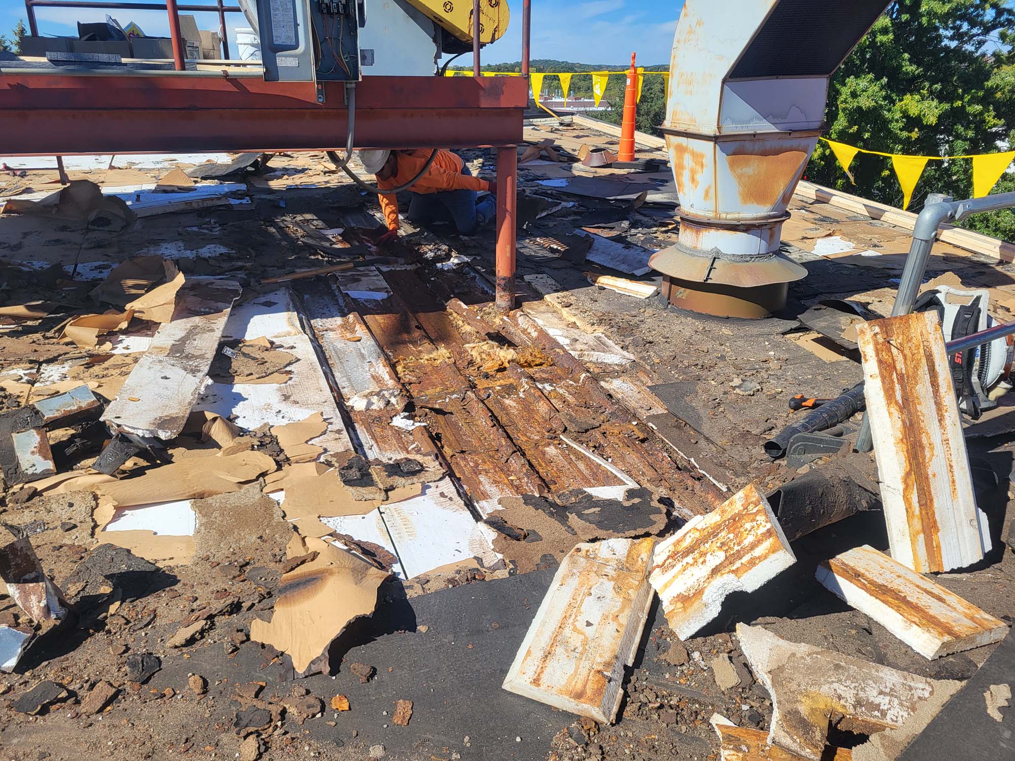 A rooftop with scattered debris and rusted metal sheets. A person in an orange shirt is working under equipment near a large vent. Yellow caution flags and trees are visible in the background.