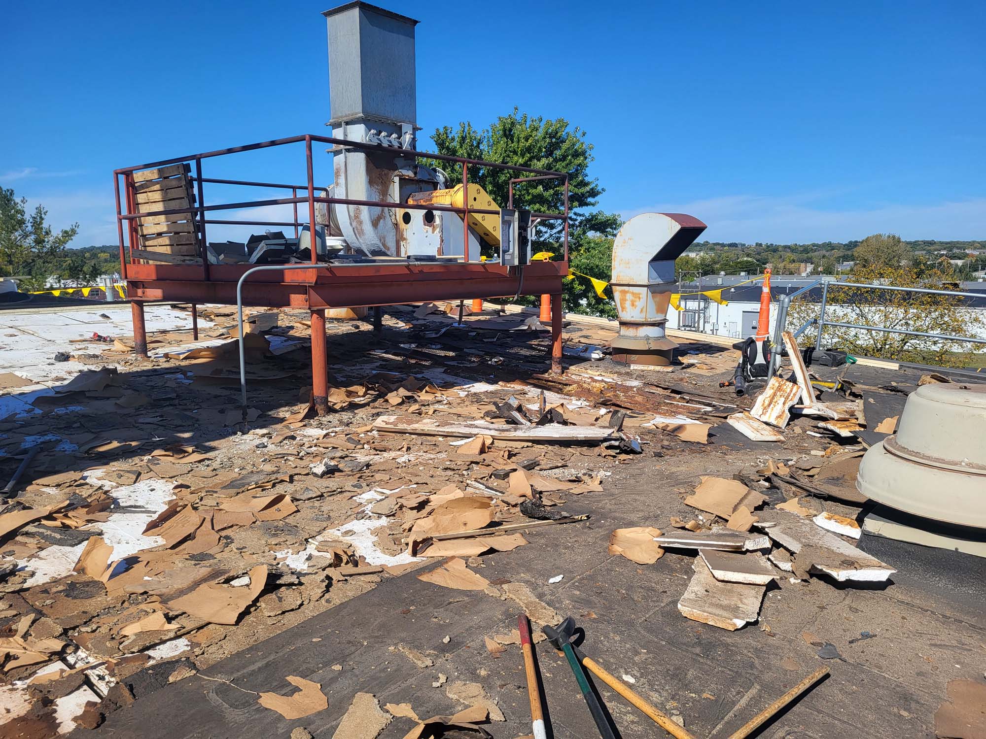 A rooftop with scattered debris, broken boards, and tools. Metal vents and a raised platform with equipment are visible under a clear blue sky, with trees and buildings in the background.