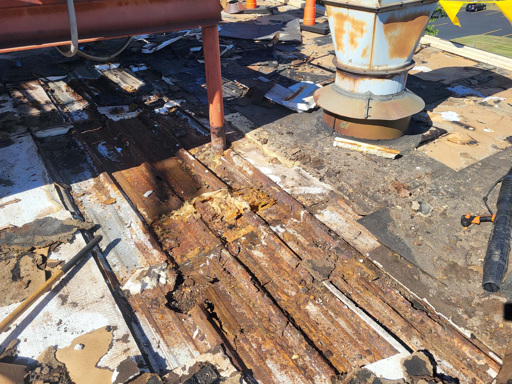 Rusty and damaged metal roofing with debris scattered around, exposed insulation, and ventilation pipes visible, indicating severe deterioration and need for repair.