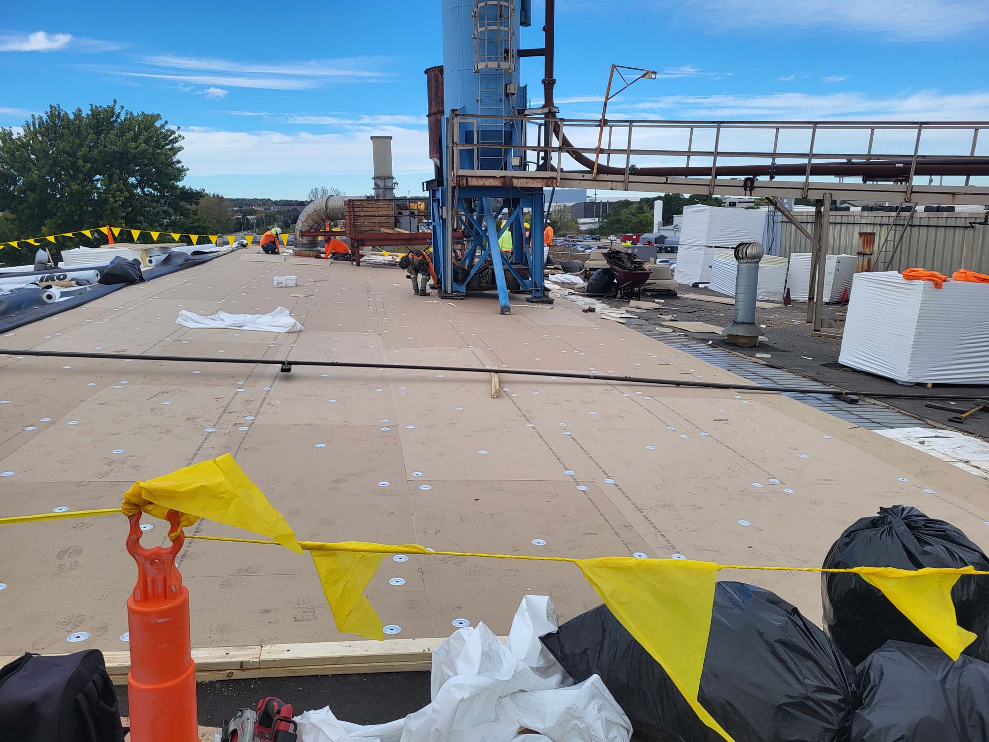 Construction workers are installing a new roof on a large building. The roof is covered with insulation boards and surrounded by yellow caution flags, with equipment and materials scattered around under a blue sky.