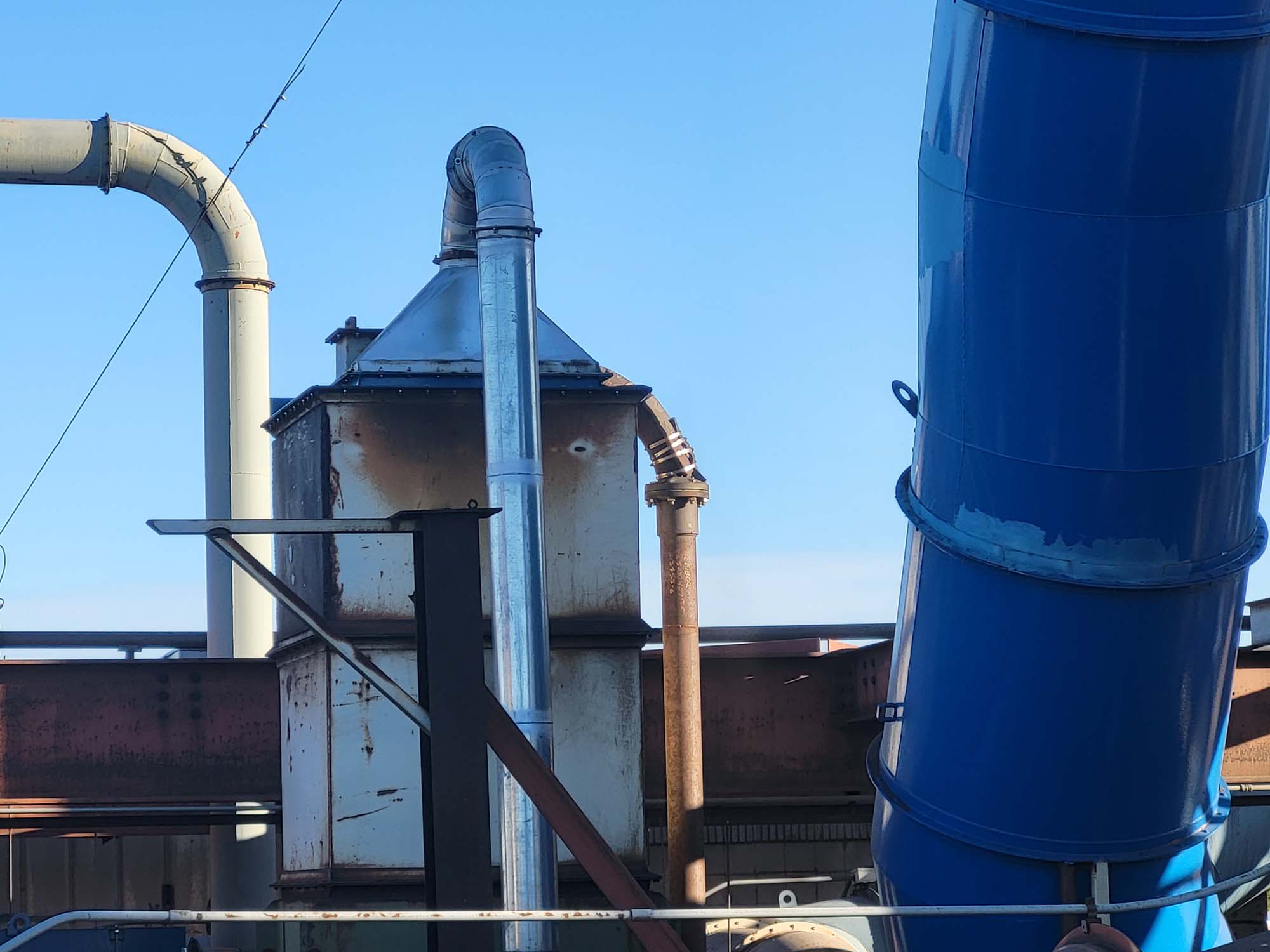 Industrial piping and metal structures, including a rusted vent and large blue duct, against a clear blue sky. The pipes are joined at different angles, showing signs of use and weathering.