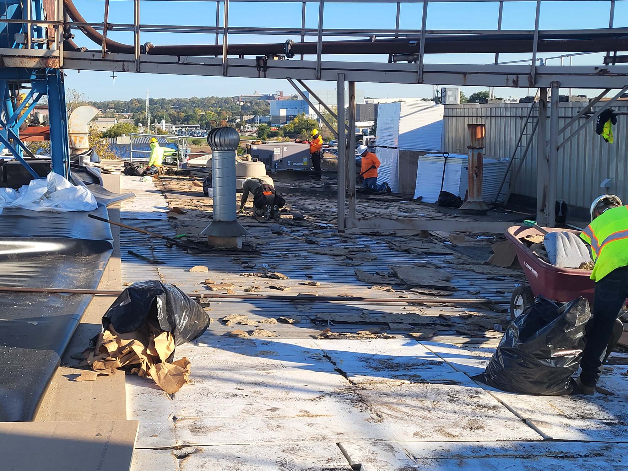 Workers in safety vests are repairing or removing debris from a rooftop under industrial pipes on a sunny day. Construction materials, trash bags, and tools are scattered across the roof. Trees and buildings are visible in the background.