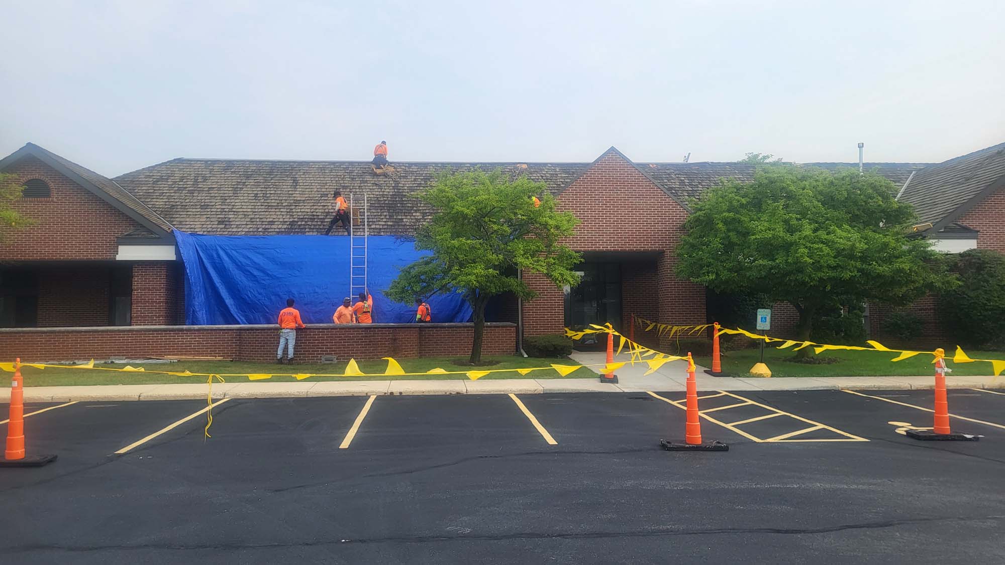Several workers in orange shirts repair the roof of a brick building, with a large blue tarp covering part of the roof. Yellow caution tape and orange cones block off the empty parking spaces in front.