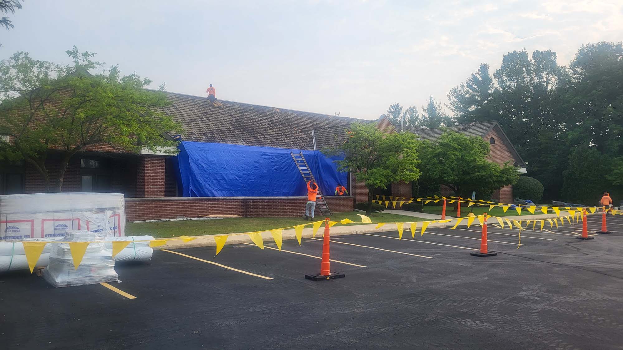 Workers in orange shirts repair a building roof covered with a blue tarp. Yellow caution tape and cones block off the area, and construction materials are stacked nearby in a parking lot. Trees surround the building.