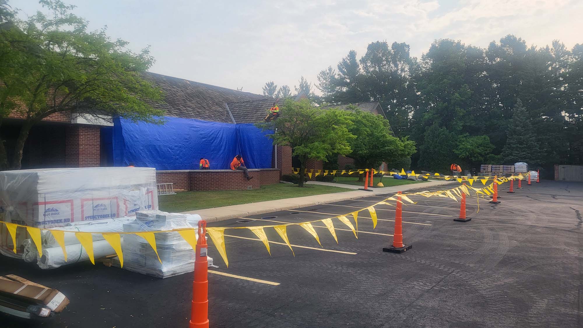 Several workers in orange vests work on a brick building covered by a large blue tarp. Yellow caution tape and cones block off nearby parking spaces, and building materials are stacked nearby. Trees line the background.