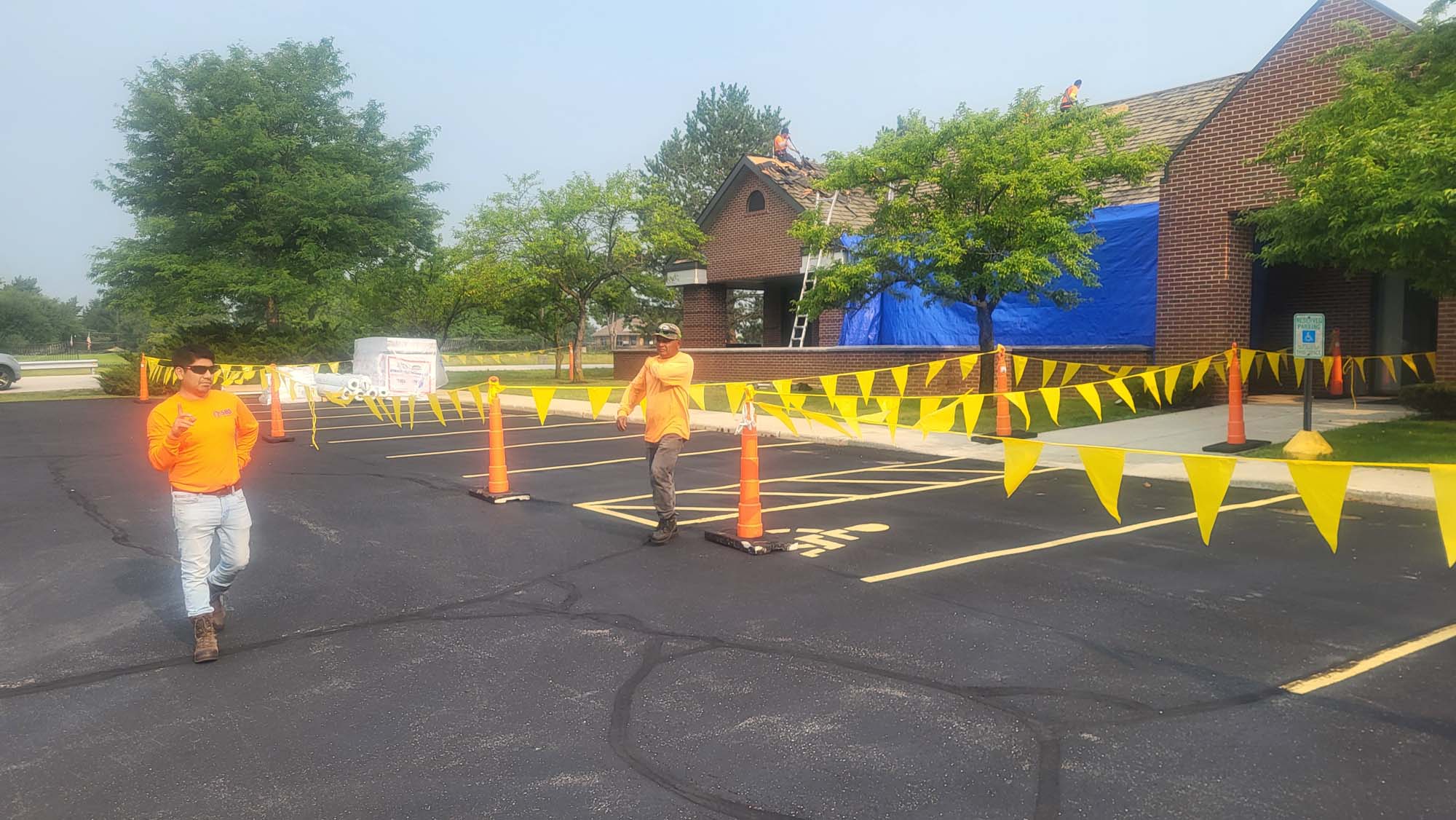 Two workers in orange shirts walk through a parking lot with yellow caution flags and cones, while roof work is being done on a brick building covered partly with blue tarp. Trees line the background.