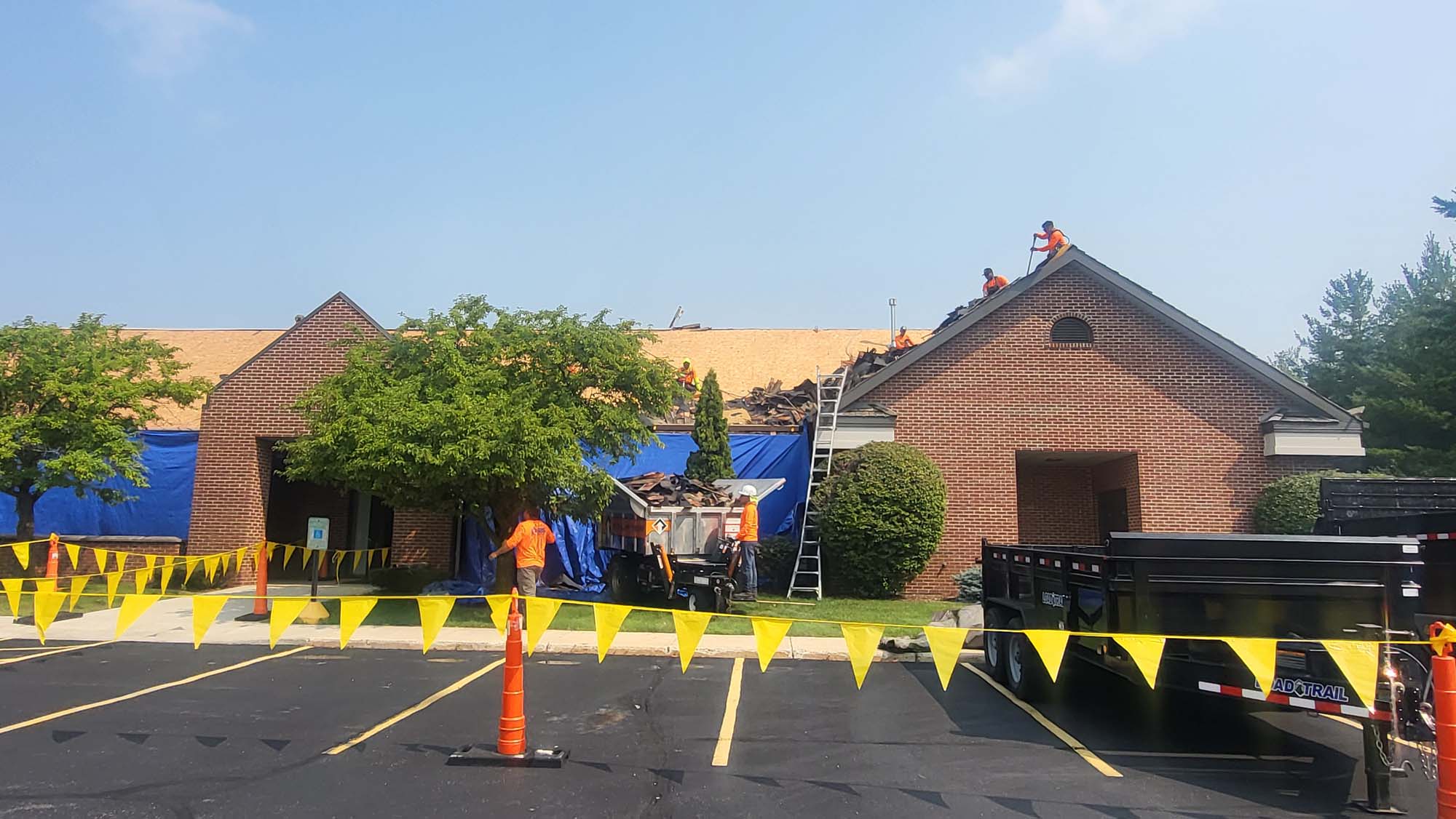 Workers are repairing the roof of a brick building, with debris being collected in a trailer below. Yellow caution tape and cones block off the parking lot area. Trees are near the building, and the sky is clear.