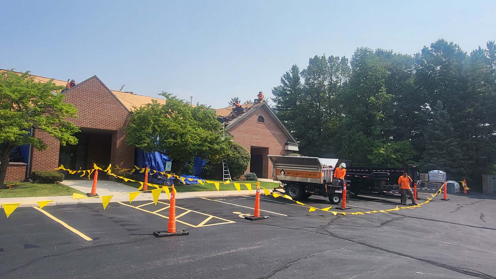 A building with workers repairing the roof, surrounded by yellow caution tape and cones. Construction materials and a truck are in the parking lot. Trees and clear sky are in the background.