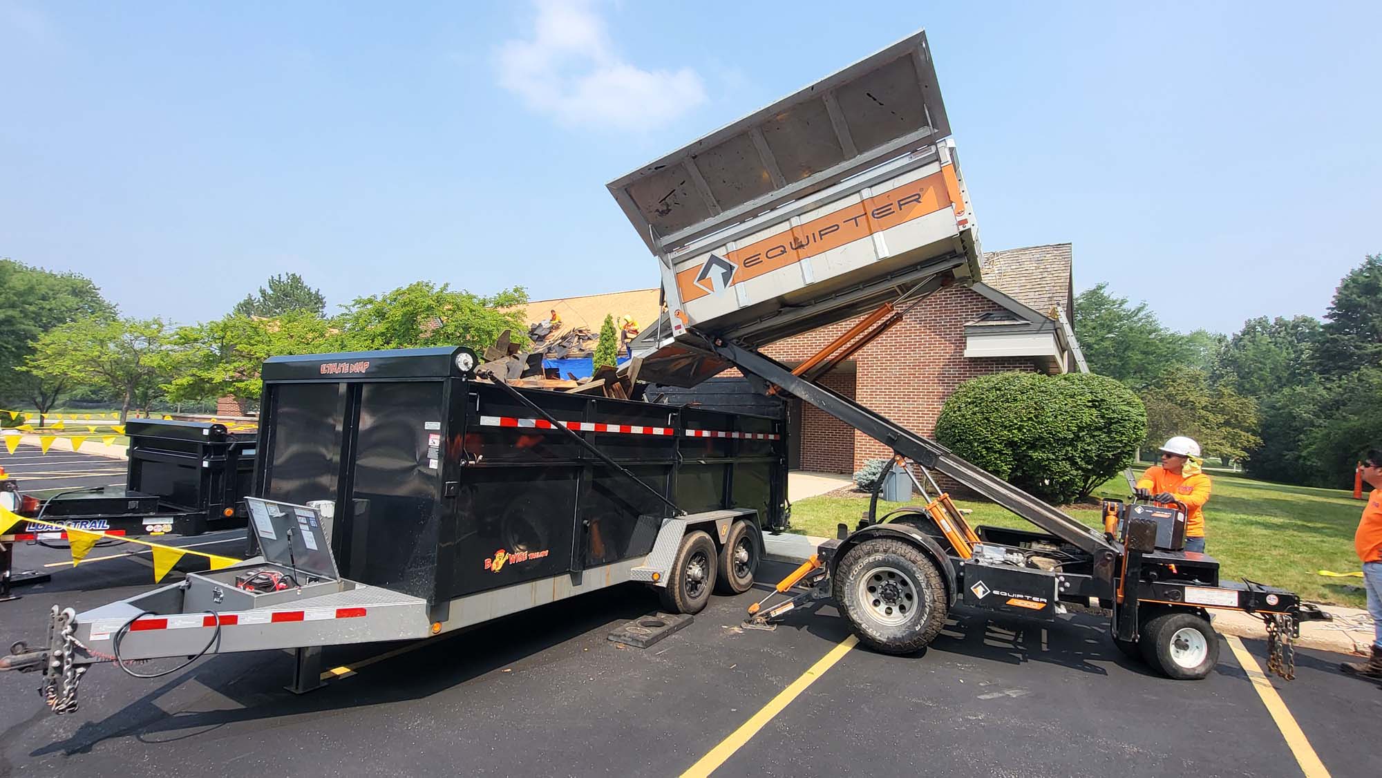 Two workers in orange shirts operate machinery to transfer debris from a raised Equipter dumpster into a larger black dumpster trailer outside a brick building on a sunny day.