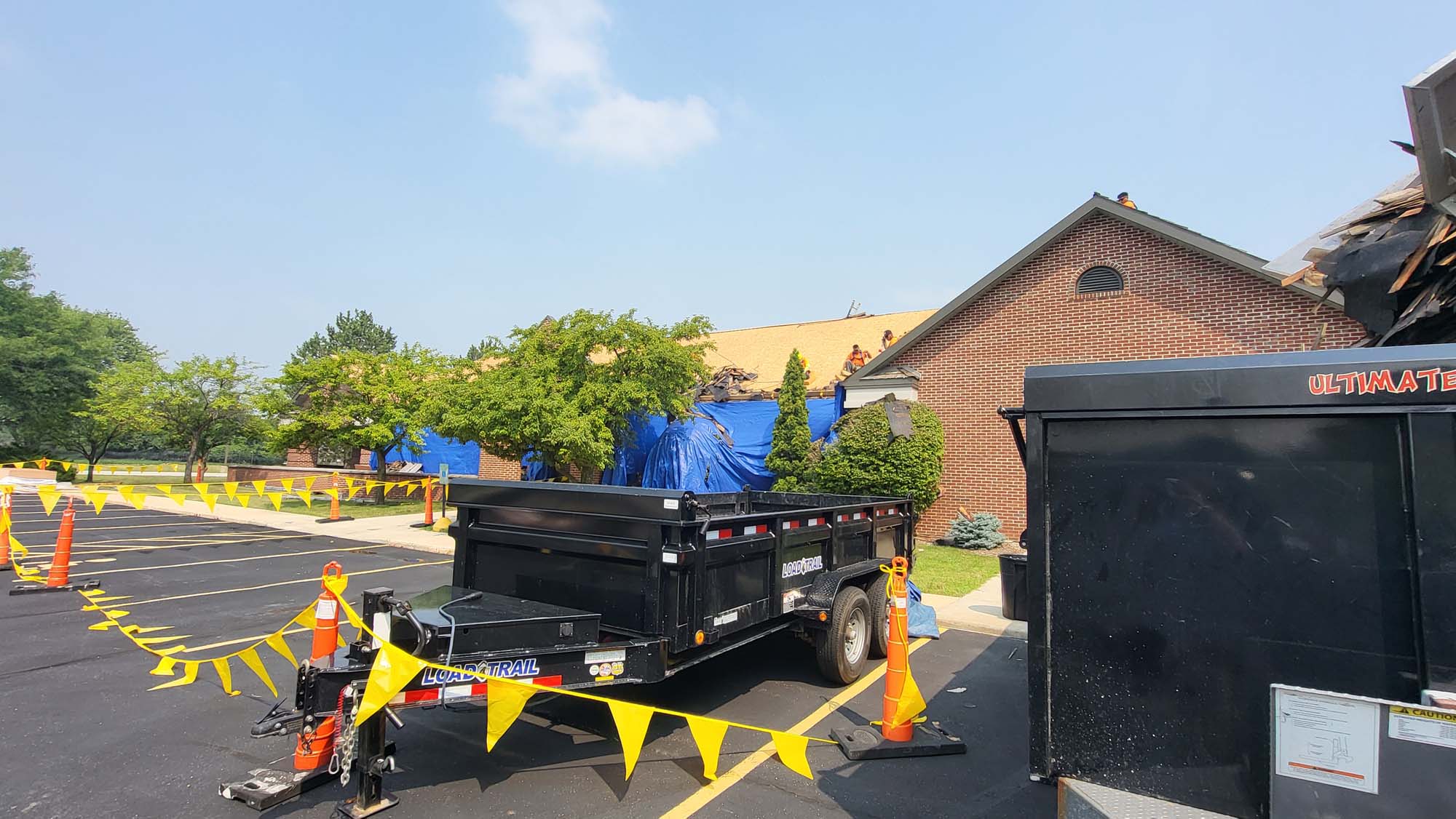 Construction site outside a brick building with workers on the roof, blue tarps covering parts of the structure, orange cones, yellow caution tape, and a black trailer in the parking lot under a sunny sky.