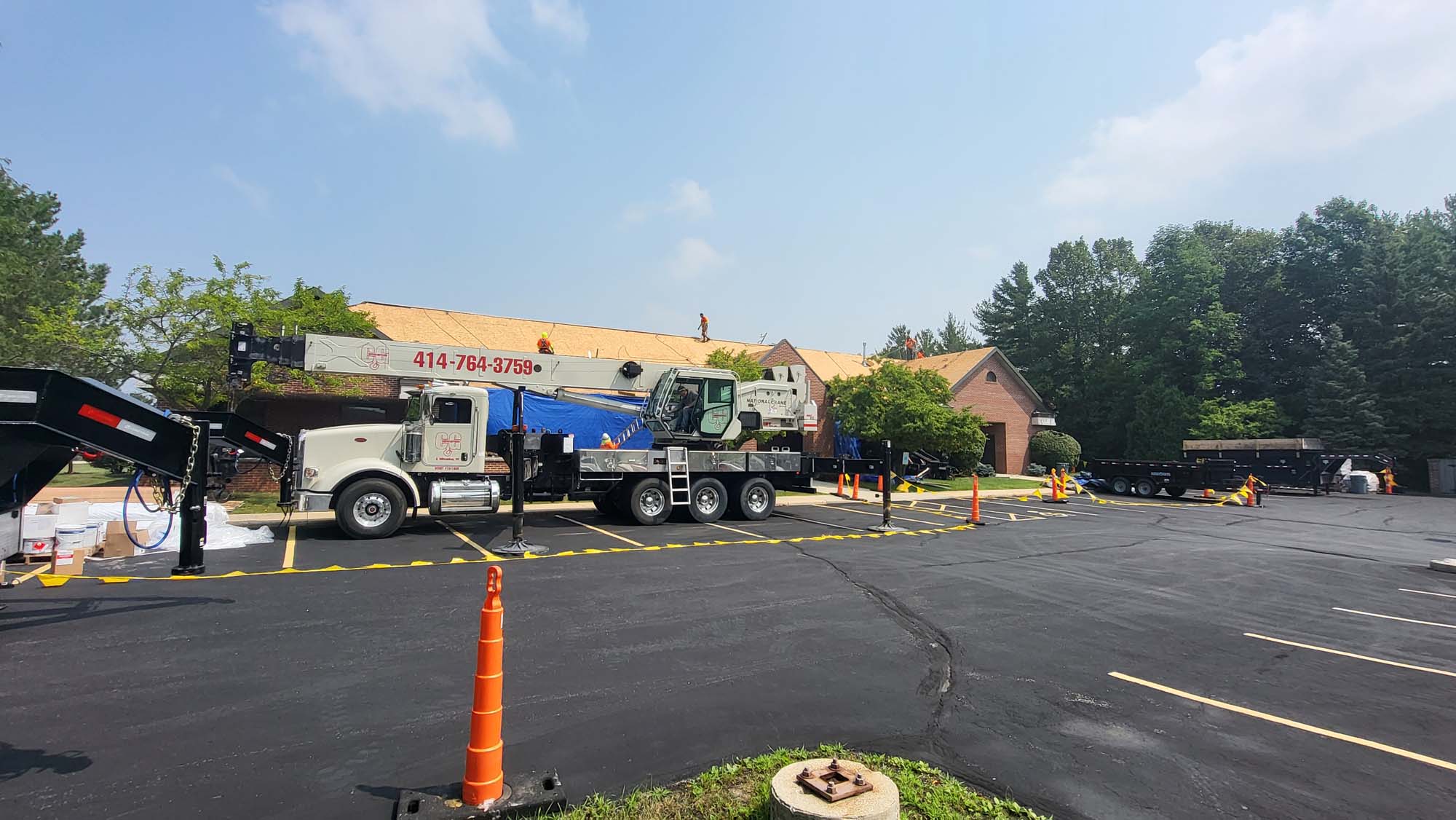A crane truck and construction equipment occupy a sectioned-off area in a parking lot near a brick building with roof work underway. Orange cones and caution tape mark the work zone under a partly cloudy sky.