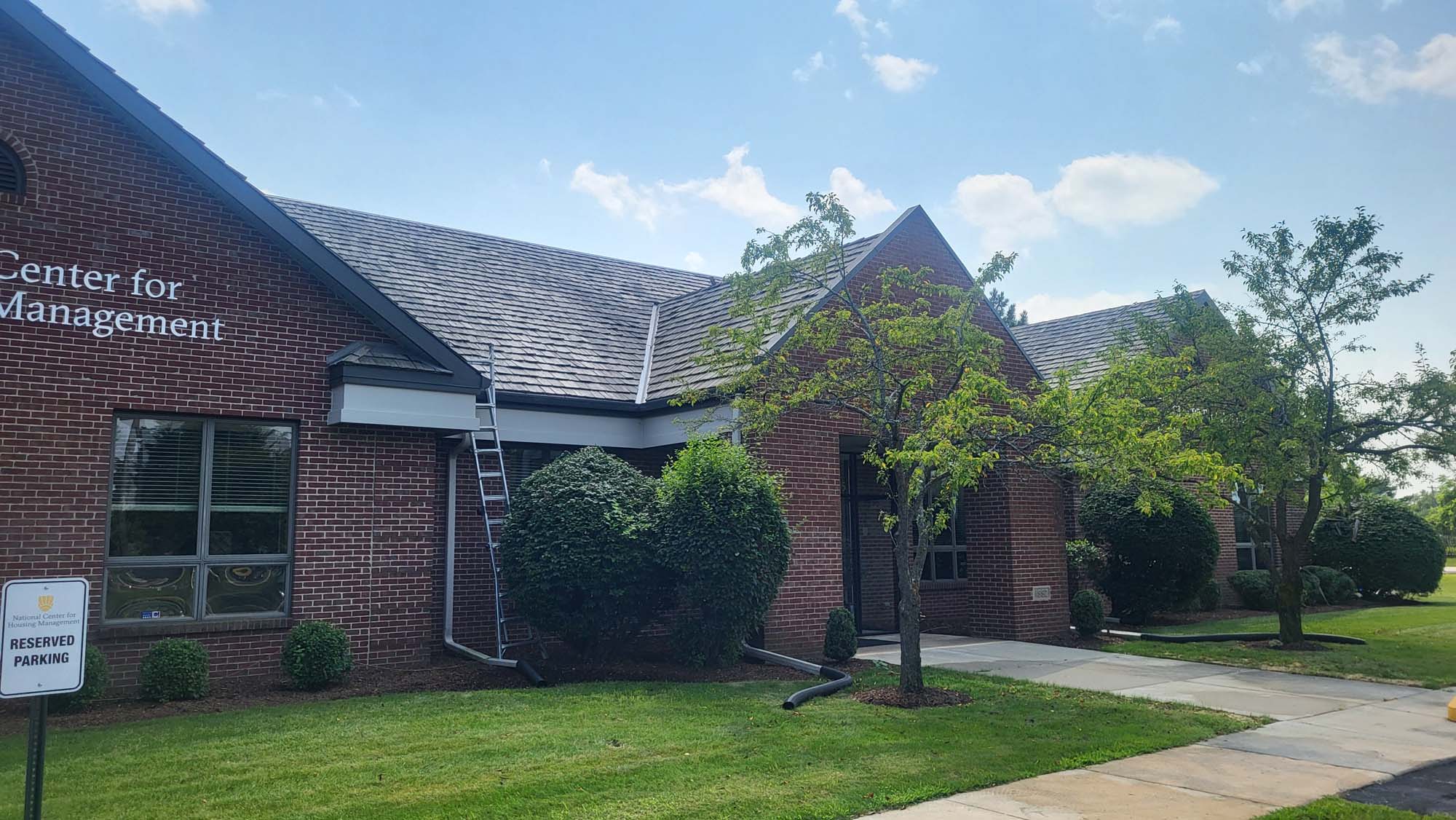 A brick building with a sign reading Center for Management, surrounded by green grass and trees. A metal ladder leans against the roof, and a Reserved Parking sign stands near the sidewalk under a blue sky.