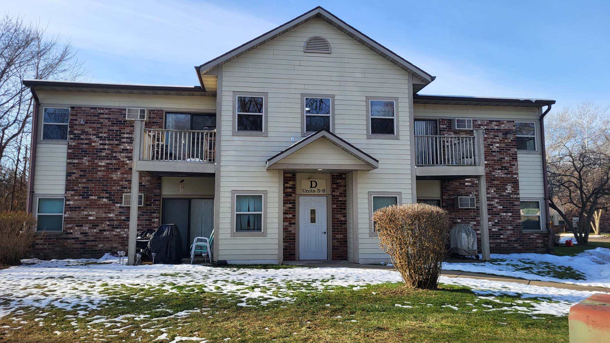 A two-story apartment building with brick and beige siding, small balconies on each side, and a door labeled D. Patchy snow covers the grass in front, with some bushes and leafless trees in the background.