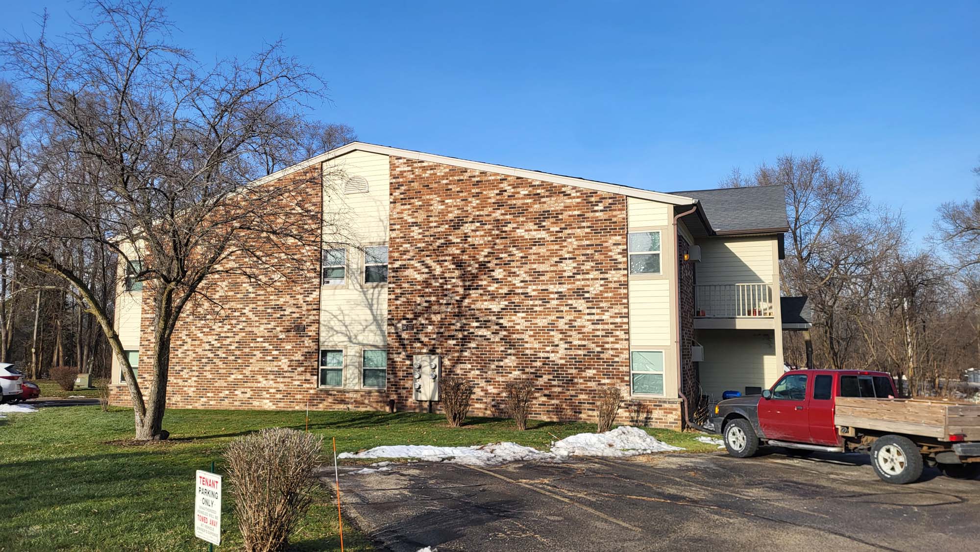 A two-story apartment building with brick and siding exterior, surrounded by leafless trees. A red pickup truck is parked nearby, and small patches of snow are visible on the grass and pavement.