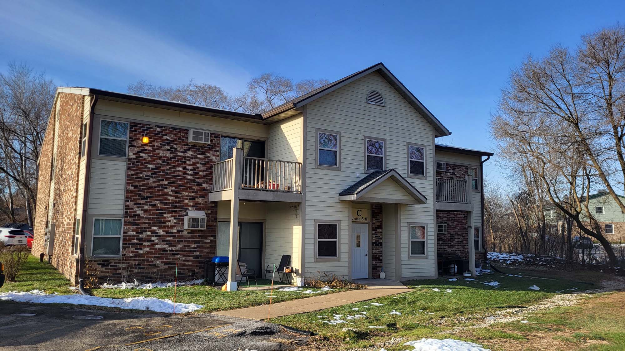 Two-story brick and siding apartment building with balconies and a central entrance, surrounded by grass, patches of melting snow, and leafless trees under a clear blue sky.