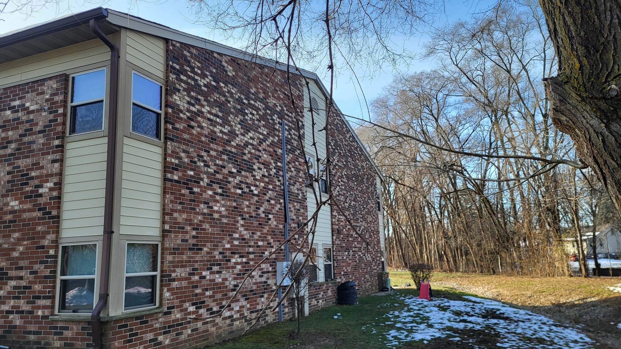 A two-story brick apartment building with tan siding stands beside a patchy lawn. Some snow is visible on the ground, and leafless trees line the background under a clear blue sky.