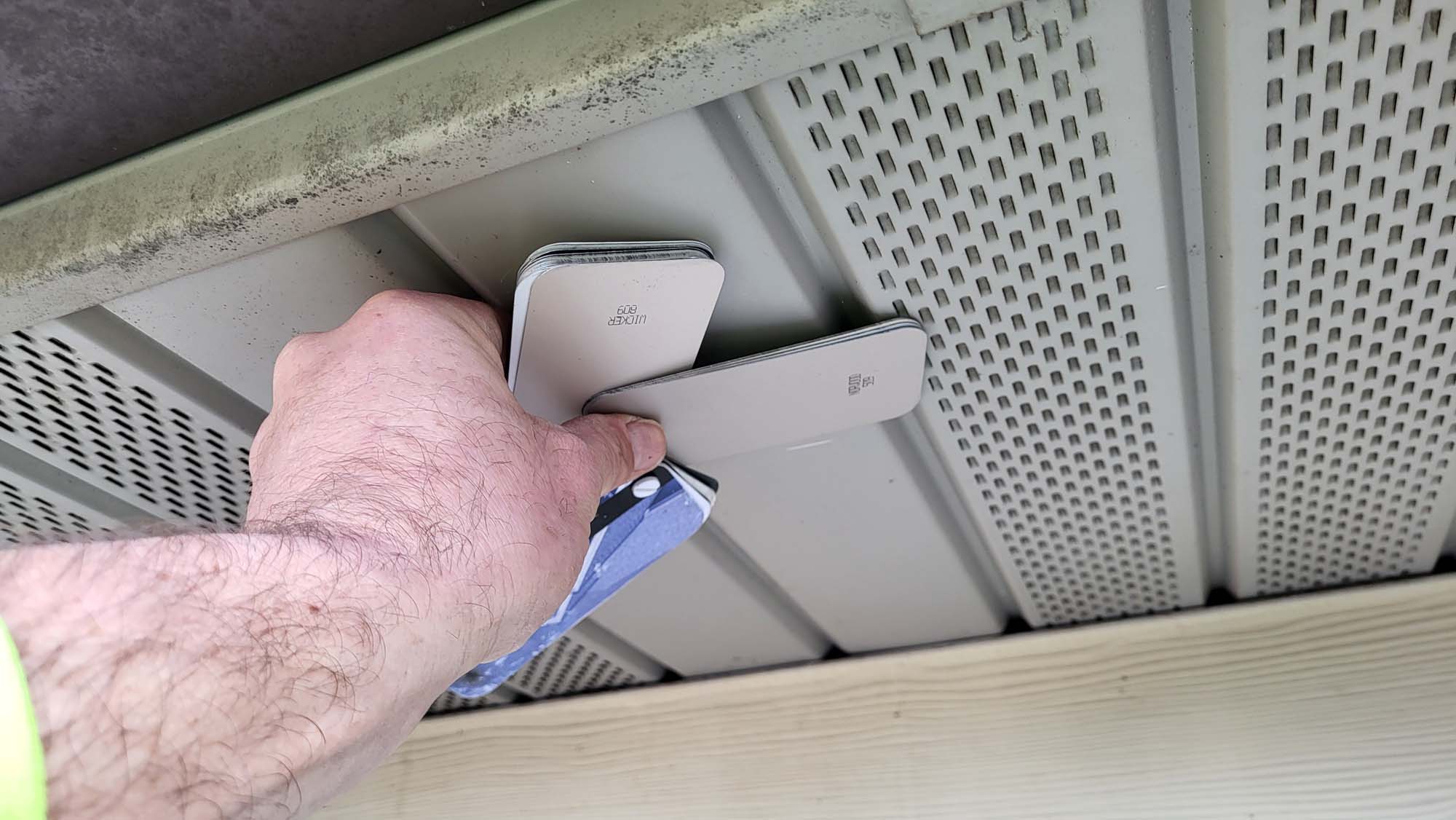 A hand holds paint swatches and a utility knife, comparing the swatches to the color of exterior house siding under a perforated soffit panel.