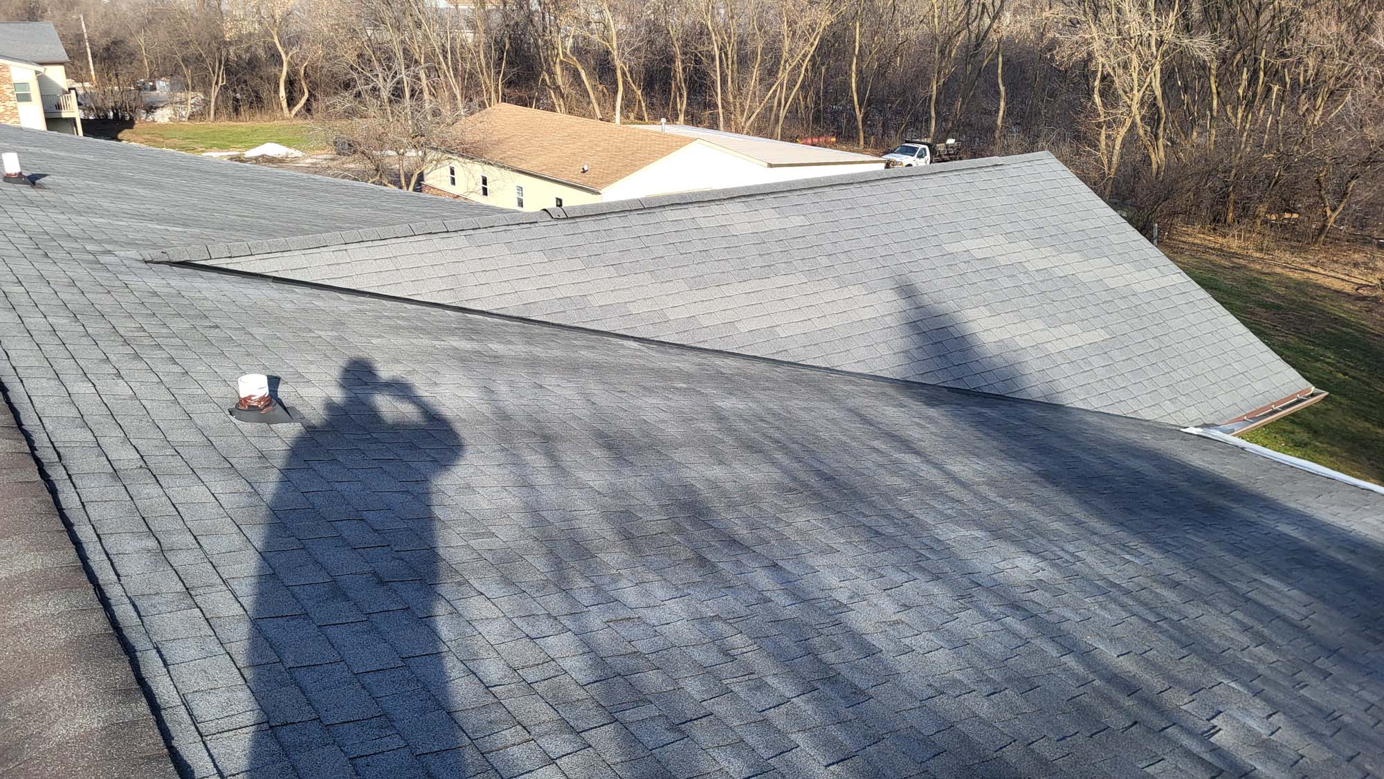 A person’s shadow is visible on a gray shingled roof, with two white roof vents and trees without leaves in the background. The photo is taken on a sunny day.