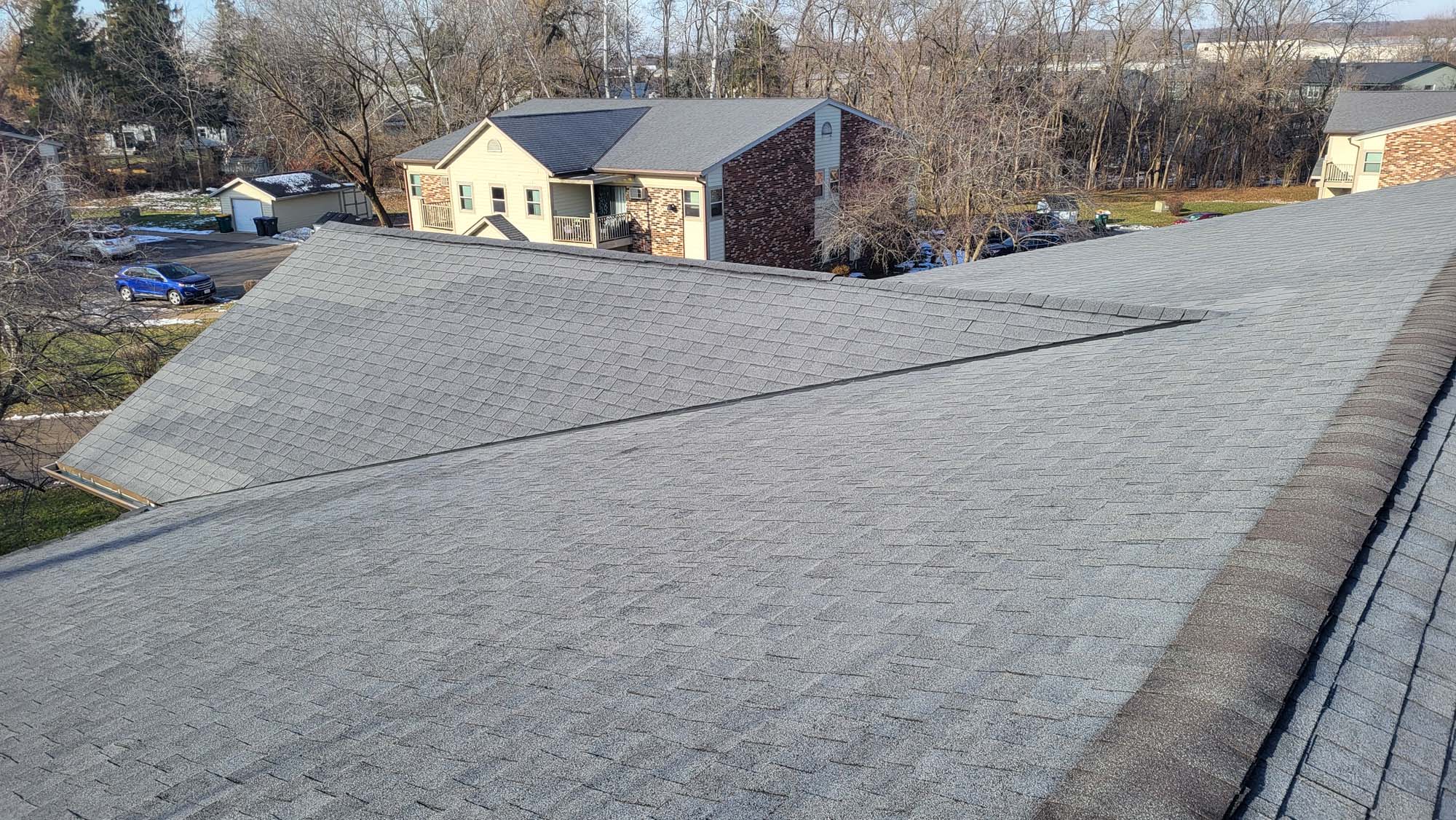 A view from the top of a grey-shingled roof, looking out towards neighboring apartment buildings and trees on a clear day, with some patches of snow visible on the ground.