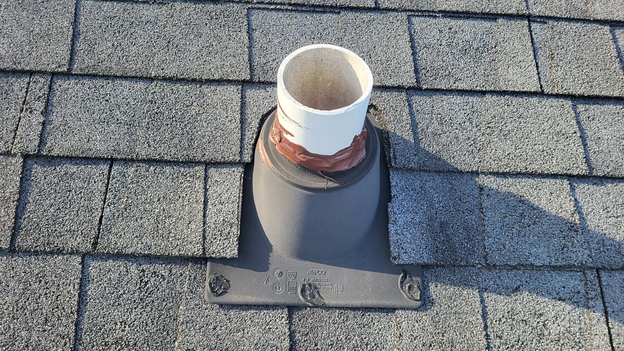 A vent pipe on a shingled roof is sealed with brown tape at the base where it meets the flashing. Some shingles around the pipe are lifted or slightly misaligned.