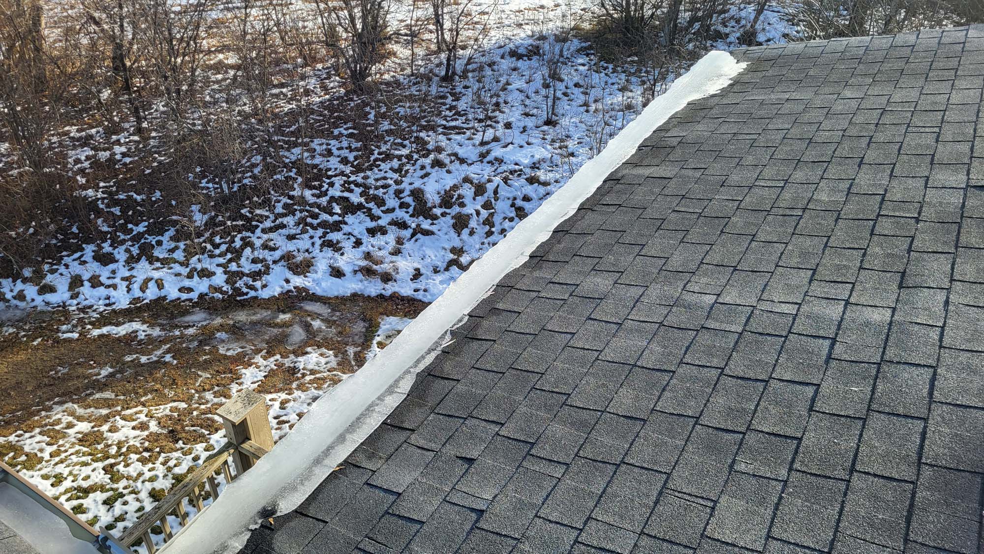 A sloped shingled roof next to a patchy, snow-covered yard with bare trees. A white line of snow remains along the roof edge, and a small section of a wooden fence is visible below.