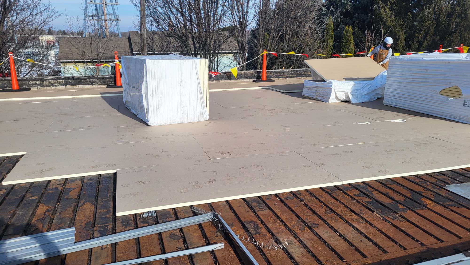 A construction worker installs large sheets of insulation on a flat rooftop. Wrapped materials and tools are nearby, and the area is marked with orange safety cones and caution tape. Trees and buildings are visible in the background.
