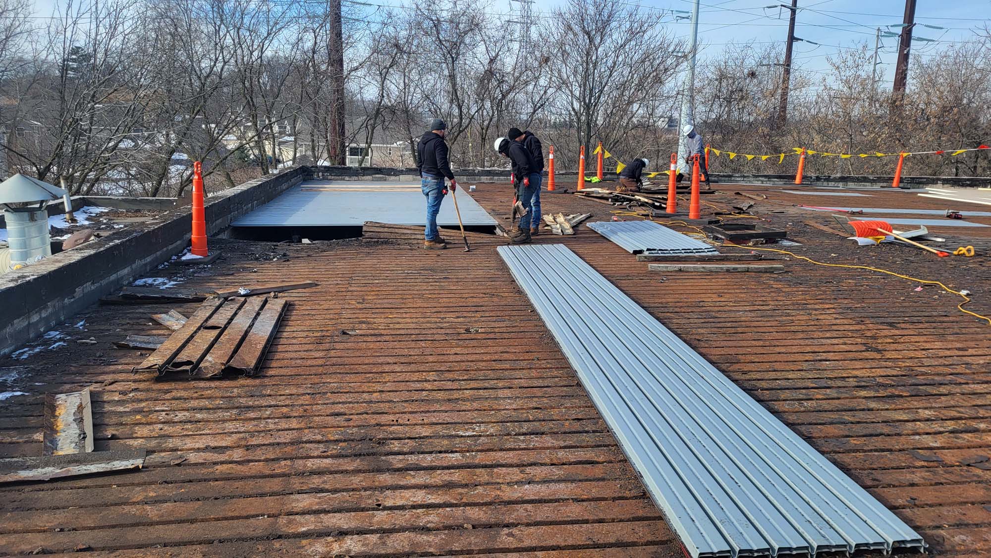 Two construction workers stand on a weathered rooftop, laying down metal sheets. Orange traffic cones, stacked materials, and tools are visible, with trees and utility poles in the background on a cold, clear day.
