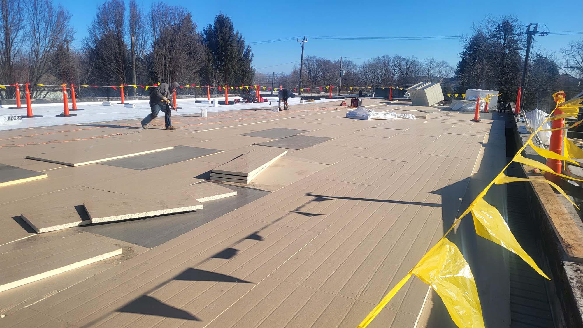 Workers walk across a flat roof under construction, with insulation panels laid out and some debris around. Yellow caution tape and orange safety barriers mark the work area. Trees and power lines are visible in the background.